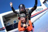 Two people skydiving tandem from an airplane. The instructor is positioned behind, guiding the diver in an orange jumpsuit. Both wear helmets and harnesses. The sky is clear and blue, and the airplane door is open, revealing the interior.