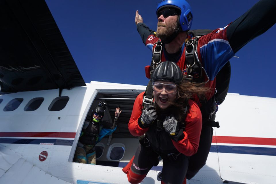 A tandem skydiving pair leaps from a plane. The man, wearing a blue helmet and dark sunglasses, holds the woman, who wears a red jumpsuit and goggles. A third skydiver waves from the plane's open door against a clear blue sky.