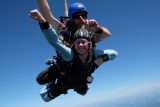 Two people are skydiving tandem in a clear blue sky. The excited participant in front smiles widely with arms outstretched, while the instructor behind points upwards, both wearing helmets and skydiving gear.