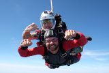 Two people tandem skydiving against a clear blue sky. The person in front is smiling widely, while the instructor behind them wears a helmet and sunglasses, both with arms extended enthusiastically.