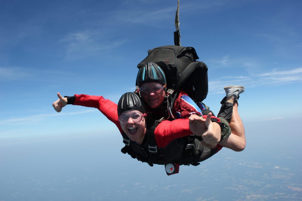 Two people tandem skydiving against a clear blue sky. Both are wearing helmets and goggles, with wide smiles and arms spread out, enjoying the thrill of the free fall. The landscape is faintly visible below them.