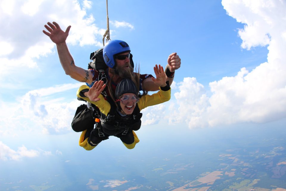 A person in a yellow suit skydives in tandem with an instructor. They are smiling broadly, wearing goggles and helmets, against a backdrop of clouds and landscape far below.