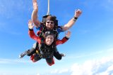 Two people tandem skydiving against a clear blue sky. The person in front wears a red jumpsuit, and both are smiling with arms spread wide. The instructor at the back has sunglasses and a black helmet. Fluffy white clouds are below them.