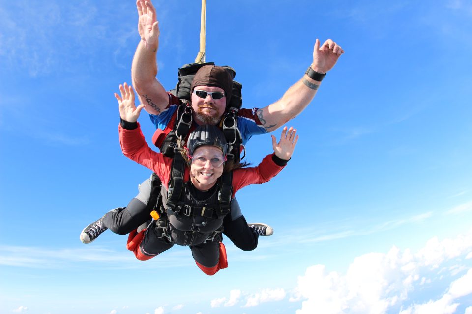 Two people tandem skydiving against a clear blue sky. The person in front wears a red jumpsuit, and both are smiling with arms spread wide. The instructor at the back has sunglasses and a black helmet. Fluffy white clouds are below them.