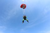 Two people tandem skydiving against a clear blue sky. They are attached to an open parachute, descending together. The main parachute is red, and one person is wearing a bright green jumpsuit.