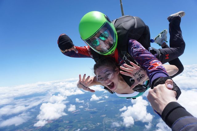 Two people skydiving in tandem against a backdrop of a clear blue sky and scattered clouds. The person in front is excitedly spreading their arms, while the instructor behind wears a green helmet. A hand is visible holding onto the instructor’s arm.