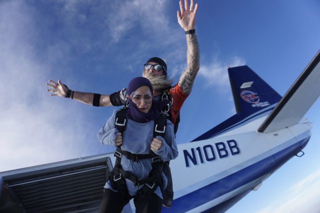 A skydiving instructor and a woman tandem jump out of an airplane. The instructor has tattoos and is wearing sunglasses, while the woman wears a hijab. They are in mid-air with the airplane visible behind them.