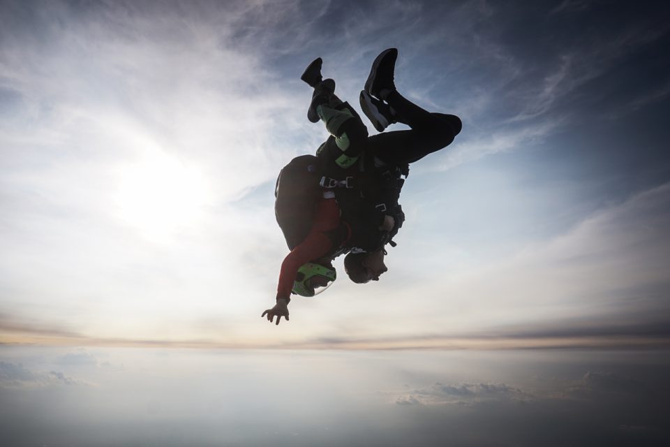 Two skydivers are in mid-air, performing a tandem dive with one person upside down. They wear helmets and harnesses against a backdrop of cloudy sky with the sun shining through.