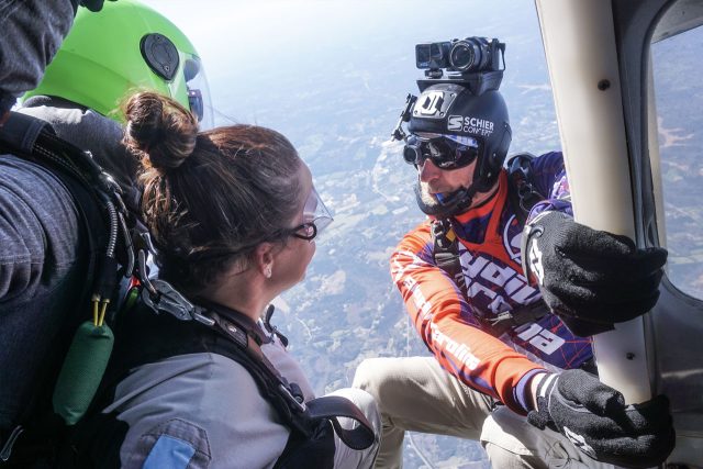 Two skydivers preparing to jump from a plane. One wears a bright green helmet, the other a helmet with a camera attached. They exchange a focused conversation, with a vast landscape visible through the open door.