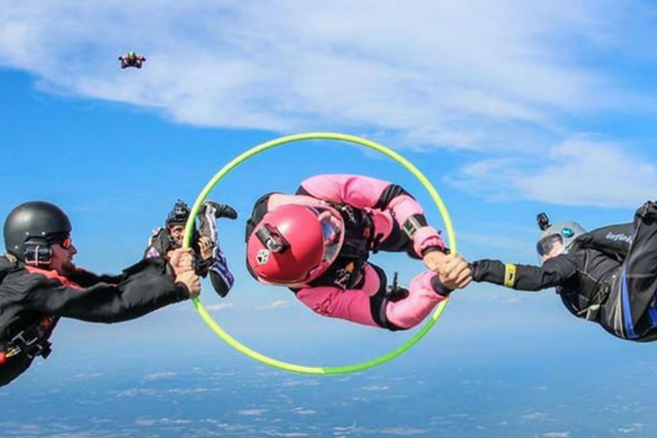 Three skydivers perform a formation in mid-air. One person is in the center wearing pink, holding a yellow hoop. They are linked by holding hands with two others wearing black and blue suits. A clear blue sky is in the background.