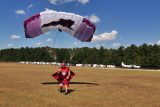 A man in a red wingsuit and helmet lands with a white and purple parachute on a grassy field. A row of trees and parked trailers are visible in the background under a blue sky with scattered clouds.