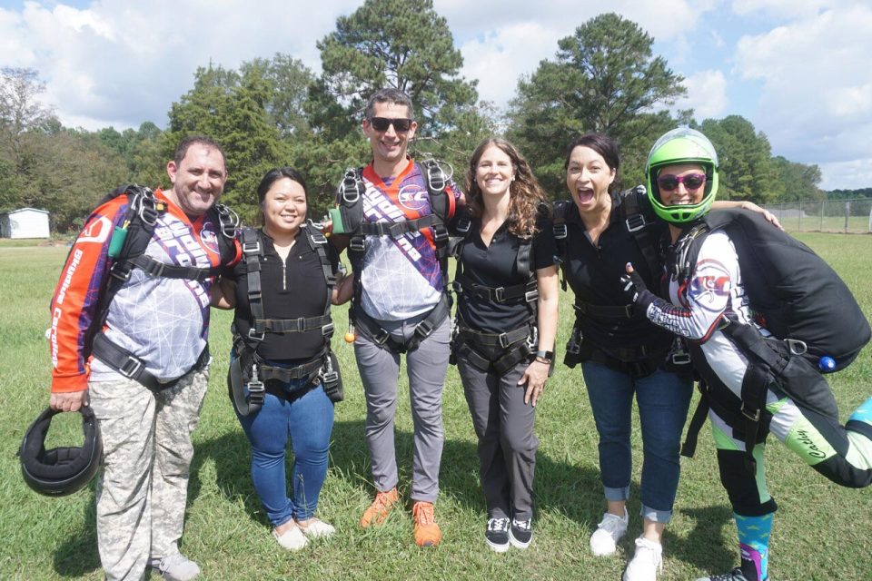 A group of six smiling people in skydiving gear stand together on a grassy field. One person is wearing a helmet. Trees and a cloudy sky are visible in the background. They appear excited and ready for an adventure.