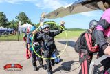 A group of people in skydiving gear prepare to board a plane. One person smiles and gestures energetically while holding a large yellow hoop. The sky is clear and the surroundings are grassy, suggesting an outdoor event.