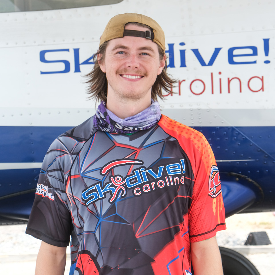 Young man smiling in front of a small aircraft. He is wearing a colorful sports jersey with "Skydive Carolina" printed on it. His baseball cap is worn backward, and he has a neck gaiter around his neck. The airplane features a partial "Skydive!" logo.