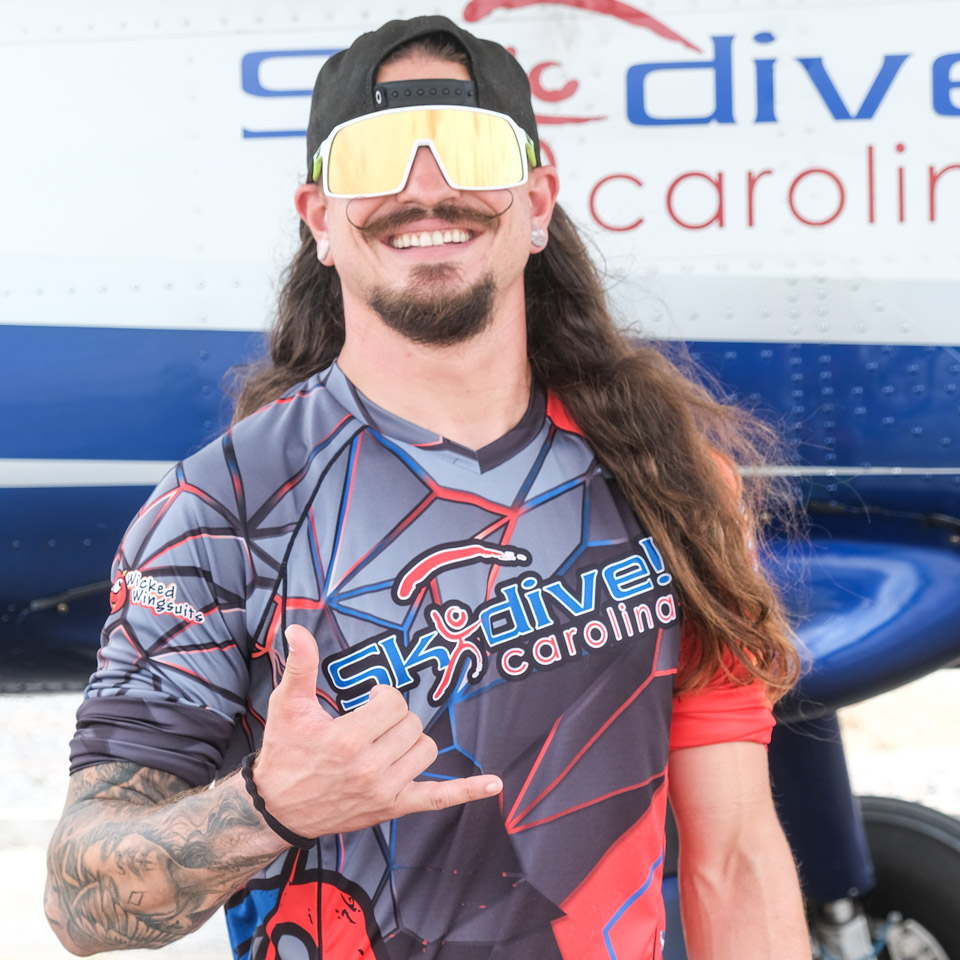 A smiling man with long hair and sunglasses stands in front of a plane. He is wearing a Skydive Carolina shirt and making a "hang loose" hand gesture.