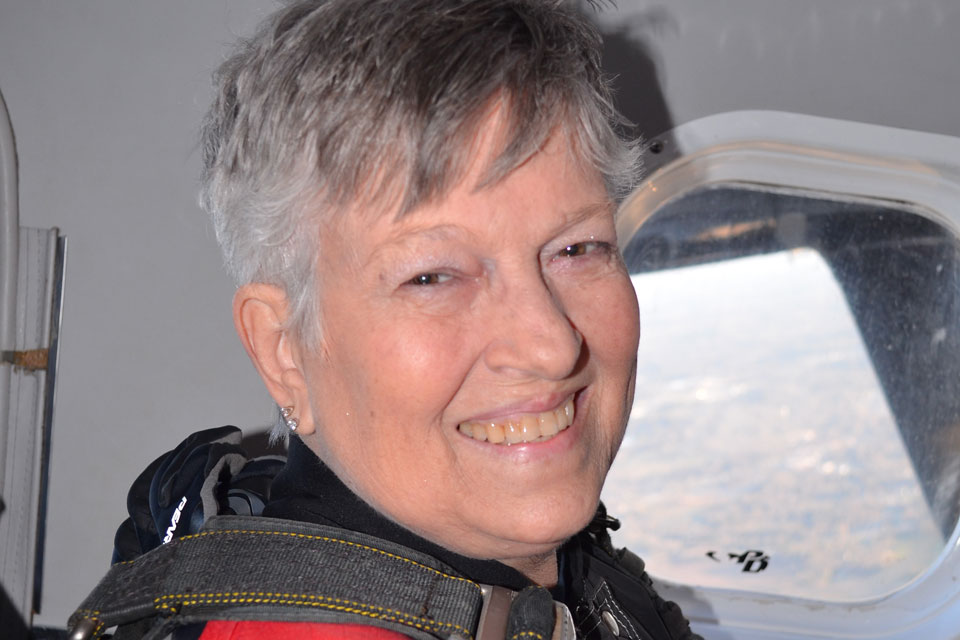 An older adult with short gray hair smiles while wearing a skydiving harness inside an aircraft. A window showing a view of the sky and landscape is in the background.