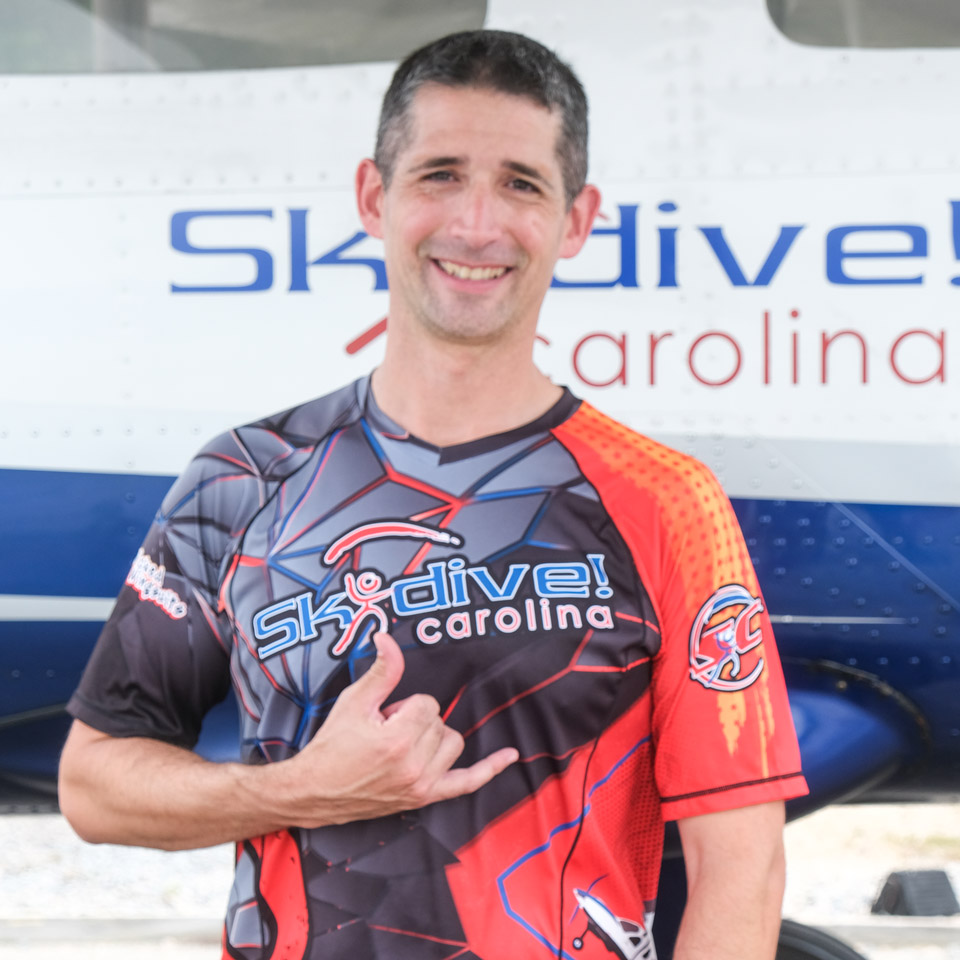 A person in a colorful Skydive Carolina shirt smiles and gestures with a hand sign in front of an airplane with the text "Skydive Carolina" on it.