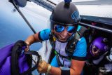 A person wearing a blue and white jumpsuit, helmet, and goggles is smiling while preparing to skydive from an airplane. Another person, also wearing gear, looks on from inside the aircraft. The sky is visible in the background.