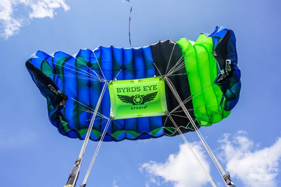 View from below of a colorful parachute with blue, green, and black sections against a blue sky. A "Byrds Eye Studio" logo is visible in the center. Fluffy white clouds are scattered in the background.