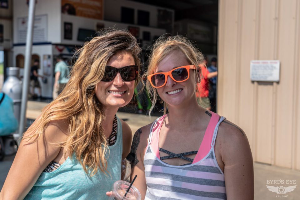 Two women wearing sunglasses stand together outside, smiling at the camera. One has long hair and wears a striped tank top, while the other has a ponytail and wears a striped shirt with bright pink sunglasses. A building is visible in the background.