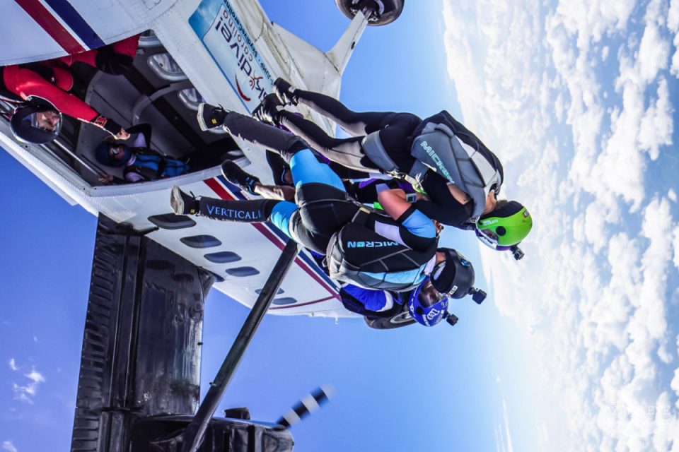 Skydivers in white and blue suits jump from an airplane into a clear blue sky. The plane is angled, with its door open. One skydiver wears a green helmet, and another holds a camera. Puffy white clouds are visible in the background.