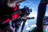 Three skydivers in helmets and gear prepare to jump from an aircraft. They are captured mid-action, looking out at the sky and clouds beneath them. One skydiver wears a red shirt, while another has green accents on their gear.