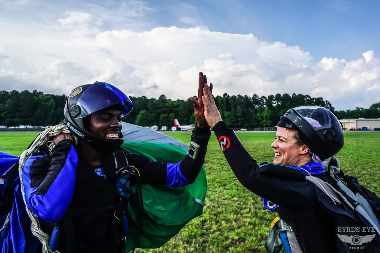 Two skydivers celebrate with a high-five on a grassy field, wearing helmets and gear. The sky is partly cloudy, and trees are visible in the background.