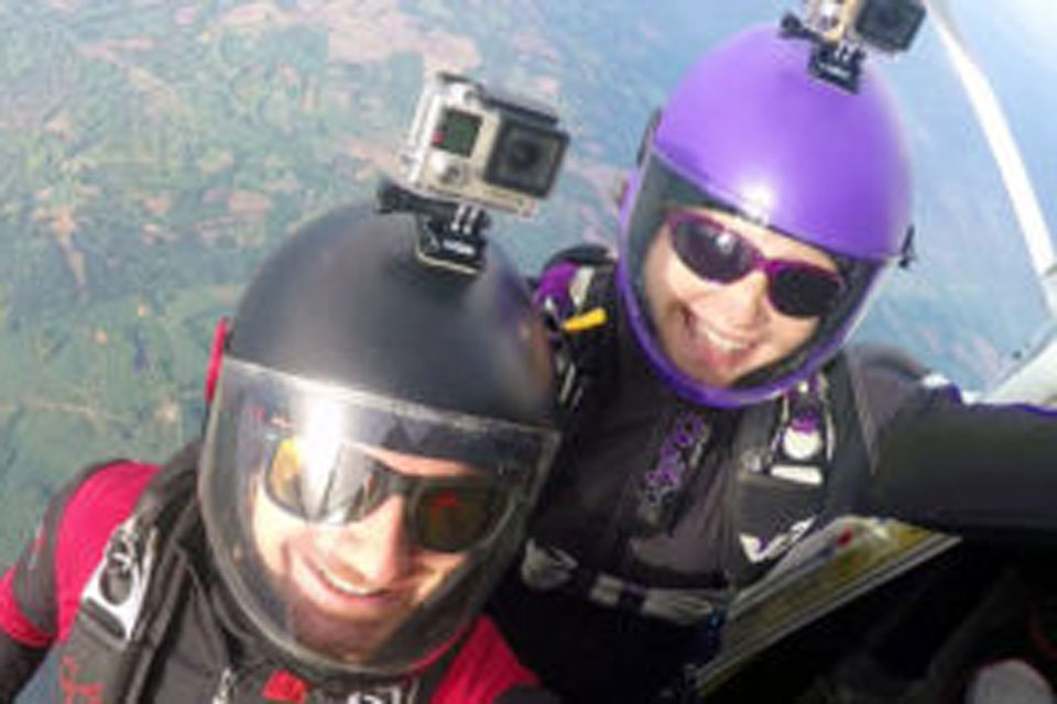 Two skydivers wearing helmets with mounted cameras and sunglasses smile at the camera. They are positioned near the open door of an aircraft, ready for a jump. The landscape below is visible in the background.