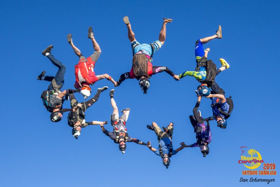 A group of nine skydivers form a circle while free-falling against a clear blue sky. Each diver is dressed in colorful gear, creating a vibrant display of human flight and teamwork.