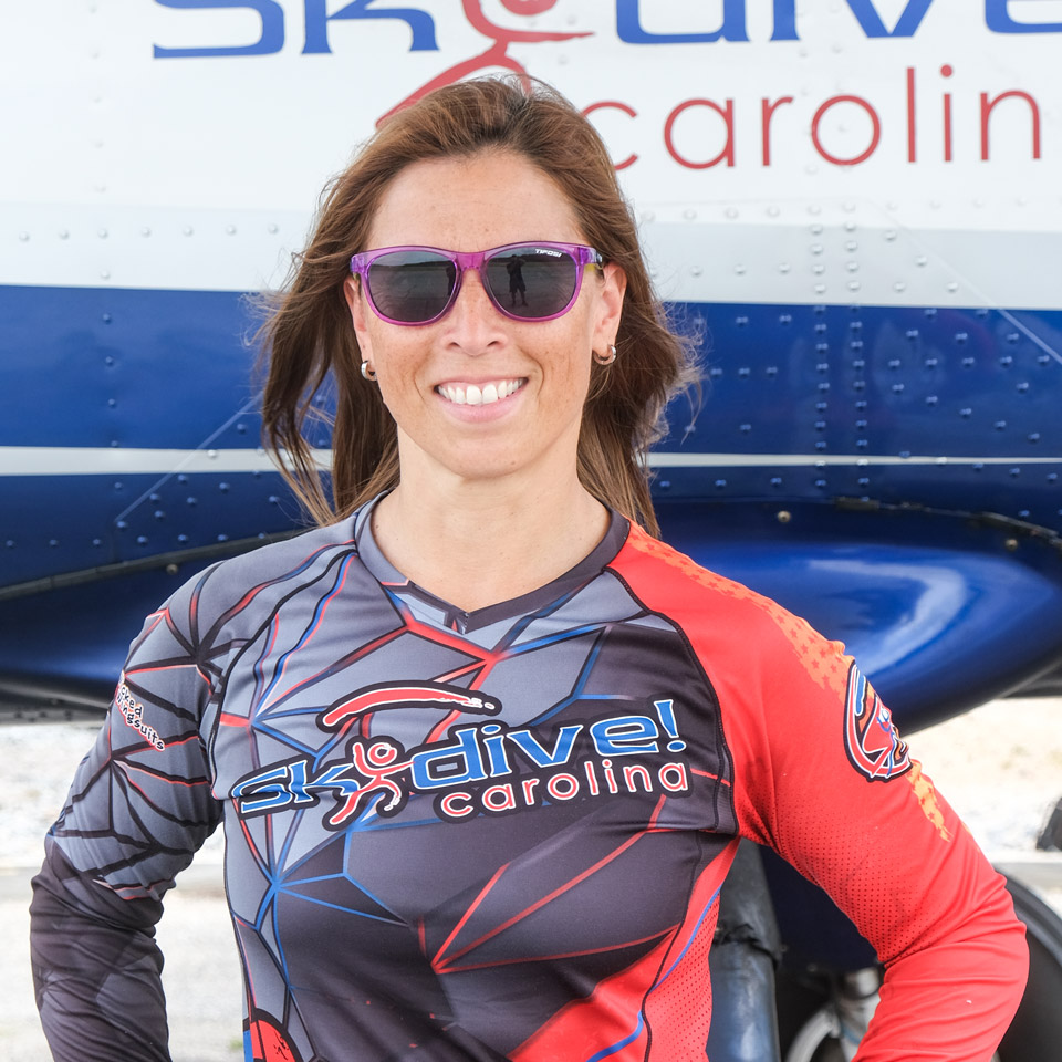 A person wearing sunglasses and a skydiving suit with "Skydive Carolina" printed on it is smiling. They are standing in front of an airplane that also has "Skydive Carolina" on it.