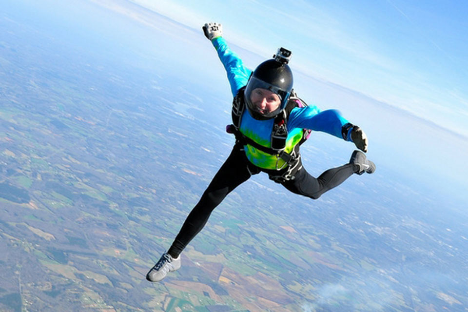 A skydiver in a bright blue and green jumpsuit is free-falling, with arms and legs extended, against a backdrop of expansive farmland and clear skies. They wear a helmet with a camera attached.