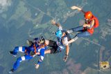 Four skydivers in colorful gear are linked together mid-air over a landscape of fields and forest. One wears an orange helmet, another blue, and two are in blue and white suits. A patch of clouds can be seen in the sky.