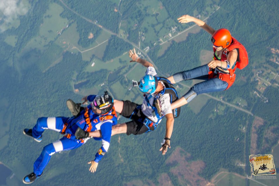 Four skydivers in colorful gear are linked together mid-air over a landscape of fields and forest. One wears an orange helmet, another blue, and two are in blue and white suits. A patch of clouds can be seen in the sky.