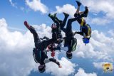 Five skydivers perform an acrobatic formation while free-falling through a bright blue sky with scattered clouds. They wear colorful jumpsuits and helmets. A view of the earth is visible far below.