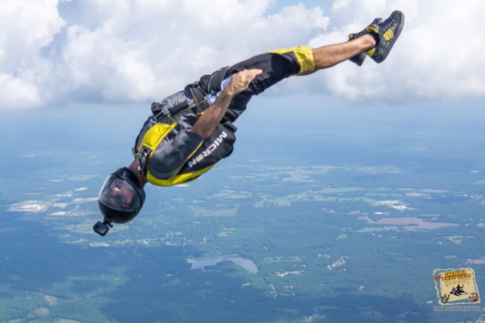 A skydiver in a yellow and black suit performs an inverted maneuver in mid-air. A camera is mounted on their helmet, capturing the expansive landscape and cloudy sky in the background.