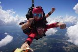 Two people are tandem skydiving, with one in a red suit and the other in a pink helmet. They are smiling and giving a thumbs-up as they fall through a clear blue sky with scattered clouds, and a landscape of fields below.