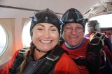 A smiling woman and man wearing skydiving gear sit inside an airplane. They have helmets and goggles on, preparing for a jump. The plane's window is visible in the background.