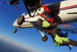 Two skydivers in bright suits jump from a white airplane against a clear blue sky. One is wearing a green suit and the other a red and blue suit. The plane's landing gear is visible, and the propellers are blurred due to motion.