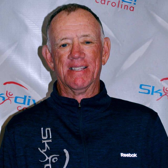 A man wearing a Reebok jacket stands in front of a backdrop with "Skydive Carolina" in blue and red text. He is smiling and has short hair.