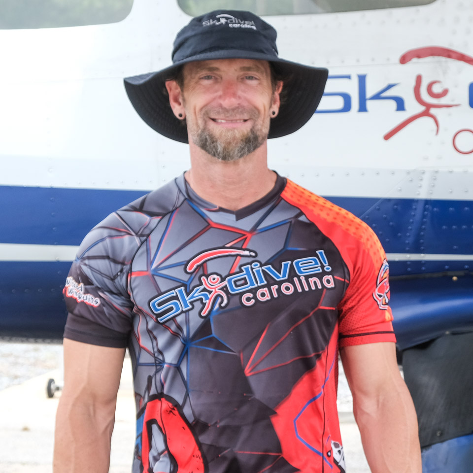 A person wearing a black bucket hat and a colorful "Skydive Carolina" shirt stands smiling in front of an airplane with partial "Skydive" branding visible on the side.