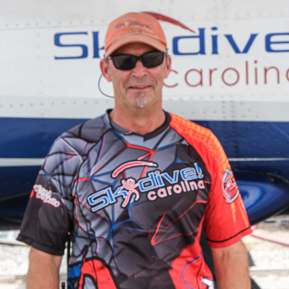 A person wearing sunglasses and a skydiving-themed shirt stands in front of a skydiving airplane. The shirt has the Skydive Carolina logo. The person appears relaxed and is smiling slightly.