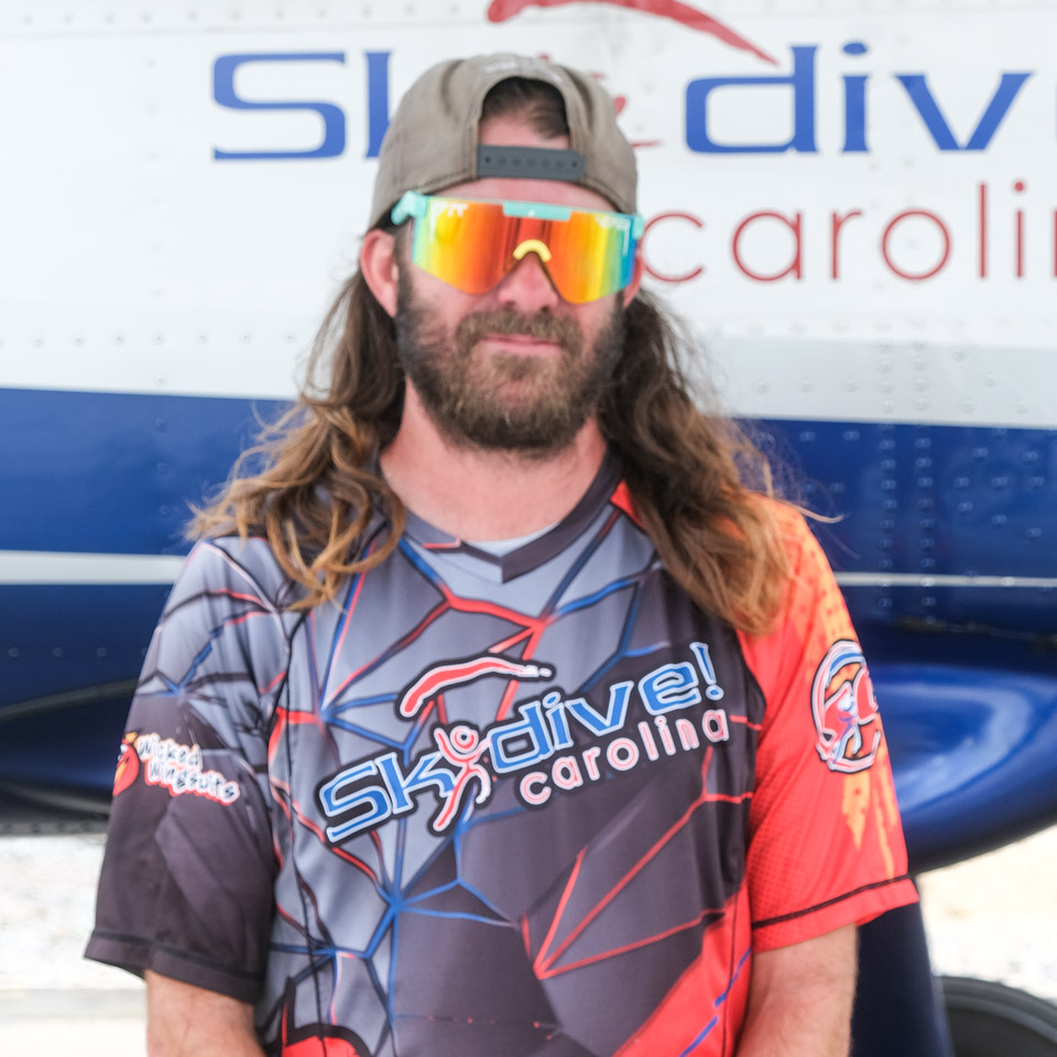 A person with long hair and a beard is wearing reflective sunglasses and a multicolored skydiving shirt that reads "Skydive Carolina." They are standing in front of an aircraft featuring the same text in partial view.