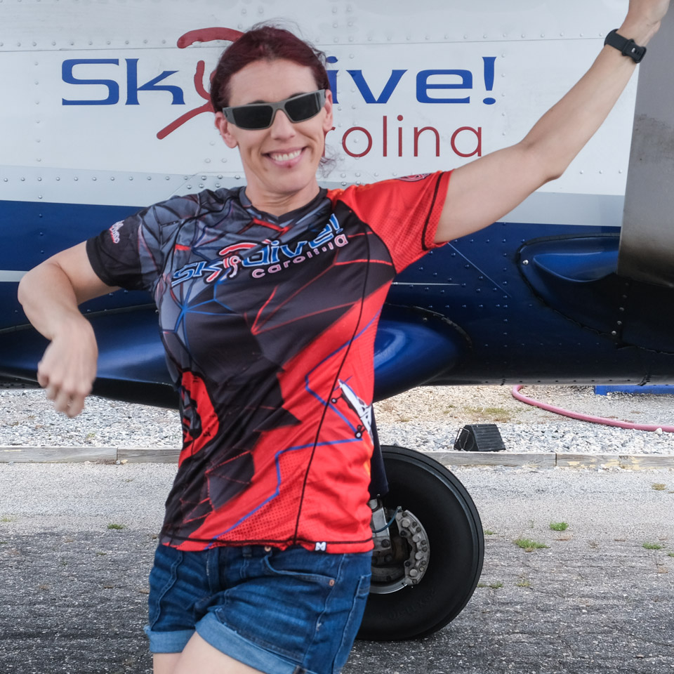 Person with red hair and sunglasses poses cheerfully in front of a propeller plane. They wear a red and black "Skydive Carolina" shirt and denim shorts, standing on a tarmac. The plane displays "Skydive!" in the background.