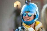 A person wearing a blue helmet with a camera mounted on top and reflective sunglasses smiles. They are dressed in skydiving gear, with a colorful scarf around their neck and a parachute harness. The background is blurred.