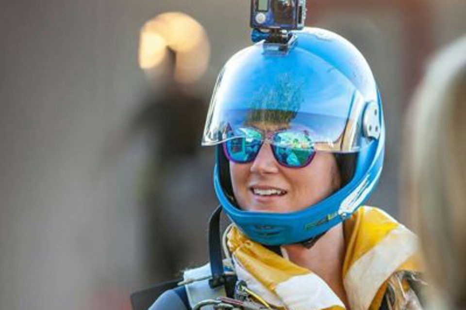 A person wearing a blue helmet with a camera mounted on top and reflective sunglasses smiles. They are dressed in skydiving gear, with a colorful scarf around their neck and a parachute harness. The background is blurred.