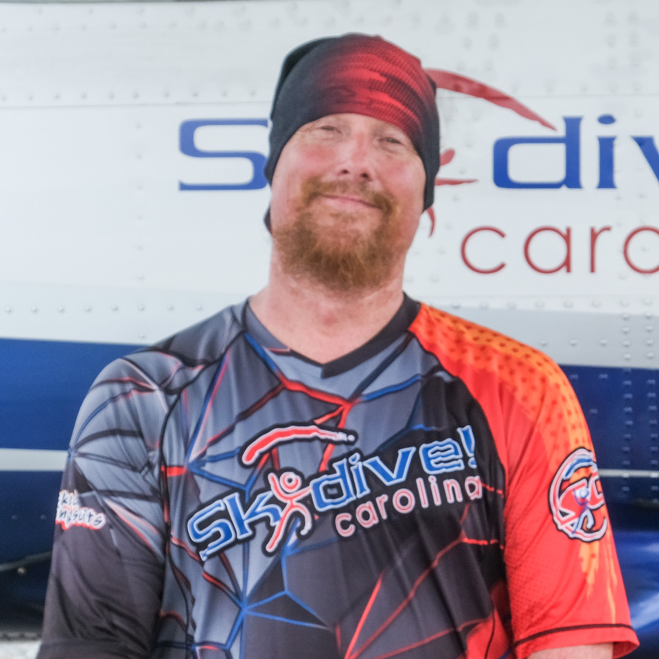 A smiling person wearing a "Skydive Carolina" shirt and beanie stands in front of an airplane. The shirt is a vibrant mix of blue, red, and white patterns. The background shows part of the airplane with partial text visible.