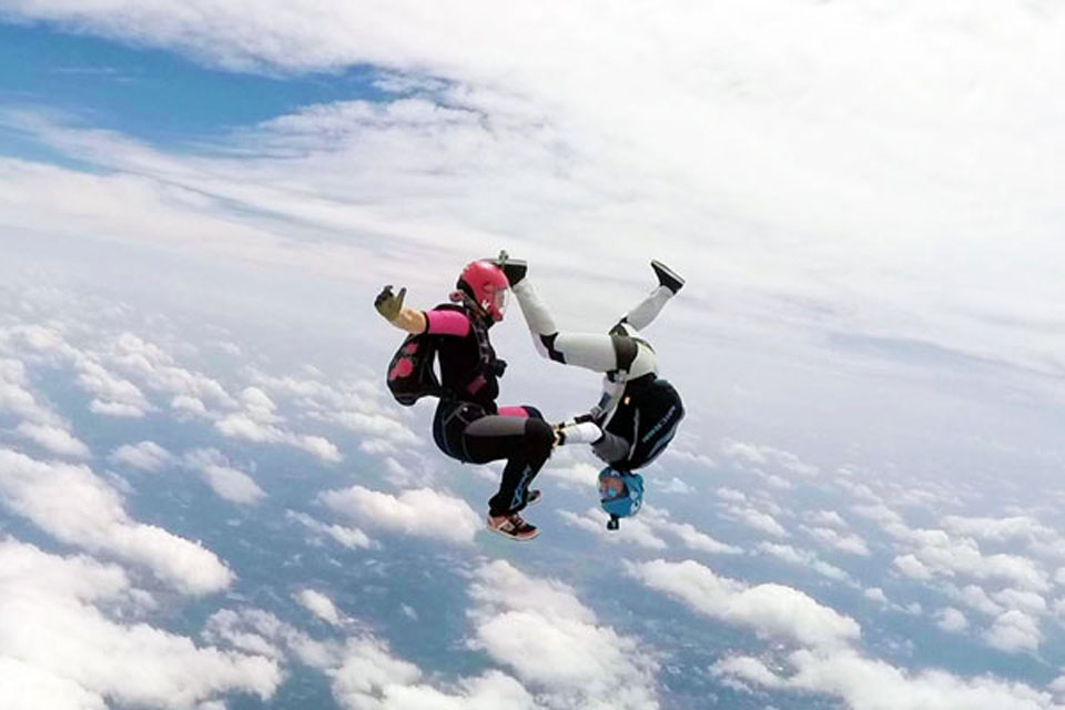 Two skydivers in colorful suits perform acrobatic maneuvers against a backdrop of clouds and blue sky. One is upright, and the other is upside-down, creating a dynamic and thrilling mid-air scene.