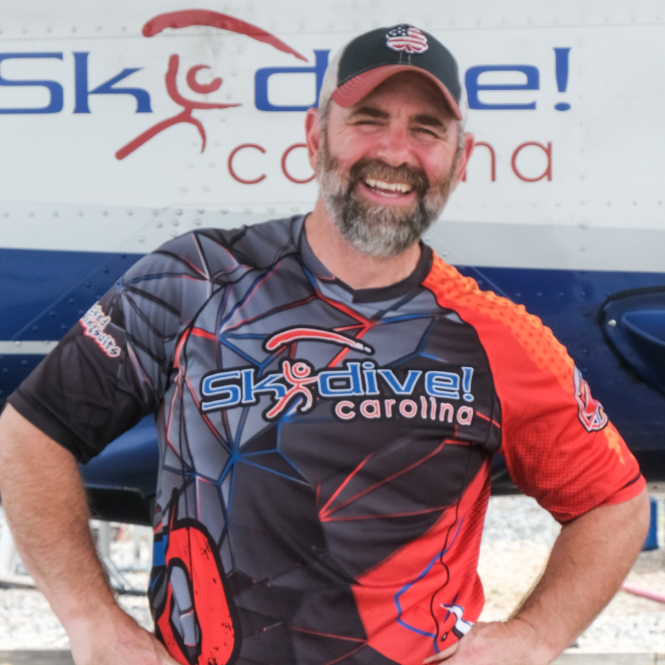 A bearded man wearing a colorful "Skydive Carolina" shirt and cap stands smiling with hands on hips in front of a plane with "Skydive Carolina" branding.