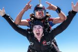 Two skydivers, in tandem, free-fall against a clear blue sky. The front skydiver, with arms spread wide, shows an excited expression, while the rear skydiver, wearing a helmet and goggles, gives a playful hand gesture.