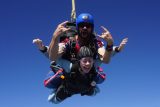 Two people are tandem skydiving against a clear blue sky. The instructor behind is gesturing excitedly with both hands while the person in front smiles broadly. Both are wearing helmets and skydiving gear.